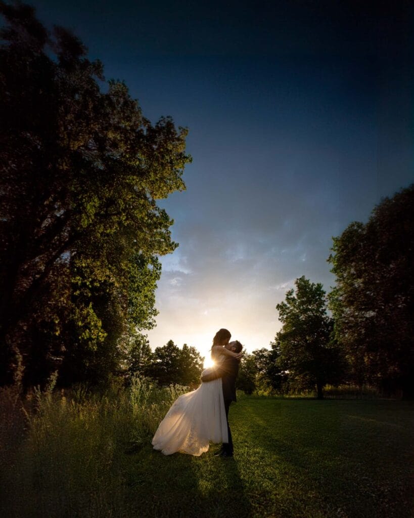 sposi durante il loro servizio fotografico di matrimonio con indosso un abito bianco e lui un abito scuro, si abbracciano con lo sfondo del tramonto e degli alberi illuminati dalla luce