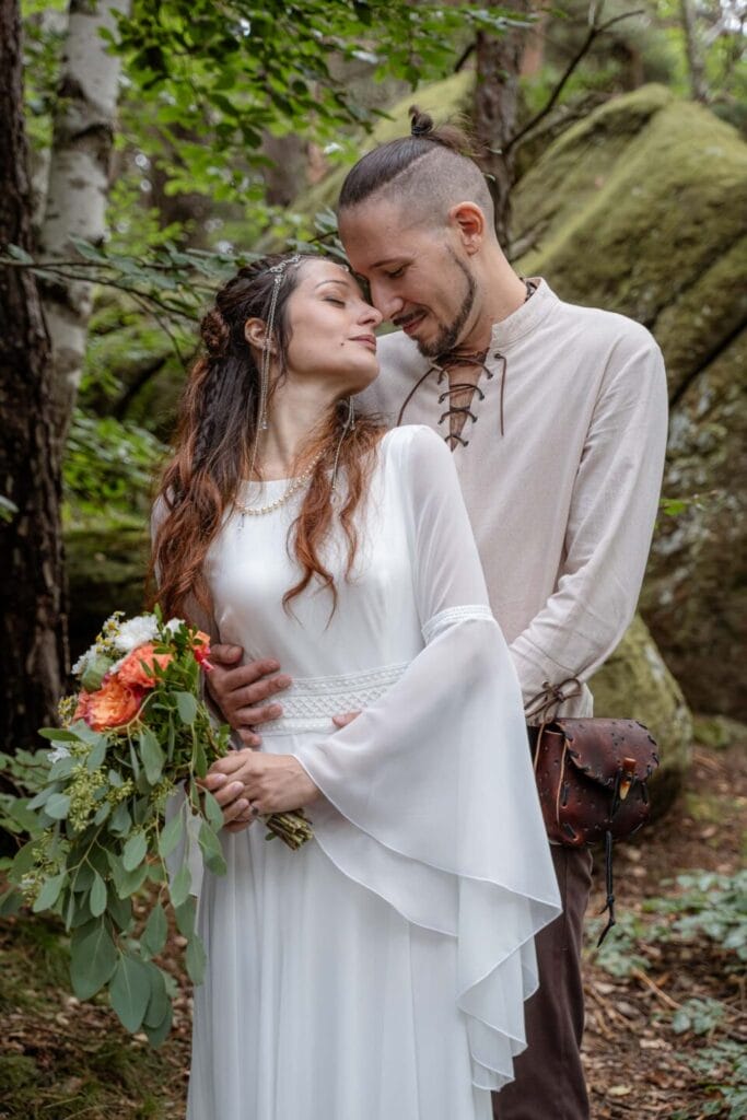 Sposi durante il loro servizio fotografico di matrimonio in stile celtico nel bosco della Val di Susa. Lei indossa un abito bianco in stile celtico e regge un mazzo di fiori in mano, lui è vestito da bardo medievale e abbraccia la sposa da dietro