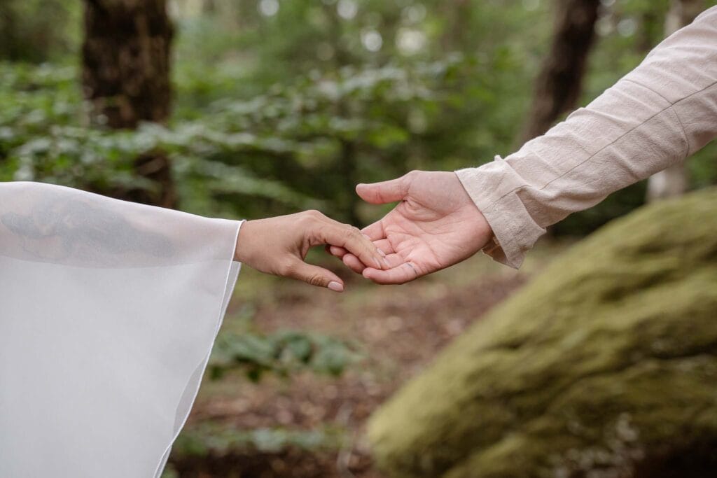 dettaglio di due mani di sposi che si incontrano a mezzaria in mezzo al bosco, lei ha una manica da abito di matrimonio celtico e lui una camicia beige