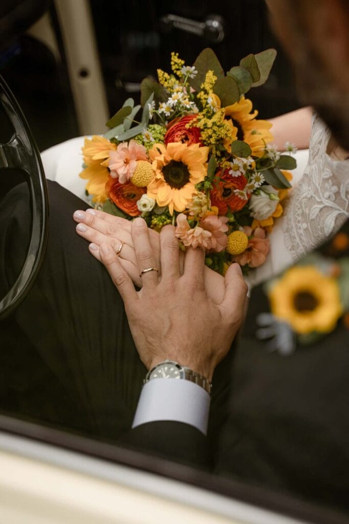 foto di dettaglio del servizio di matrimonio con due mani degli sposi di cui si vedono le fedi nuziali e il bouquet di girasoli della sposa accanto al volante di un'auto d'epoca