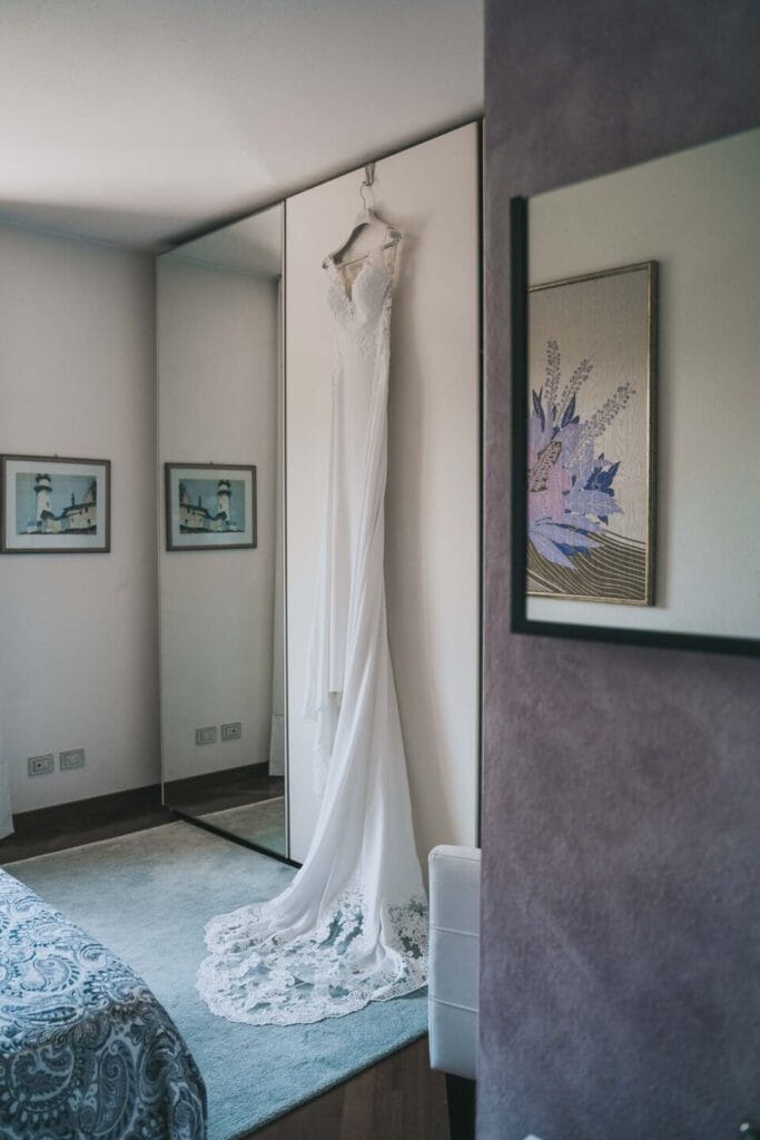 foto di un abito da sposa appeso all'armadio durante il servizio fotografico di matrimonio, l'abito è liscio e bianco e cade con lo strascico a terra, dietro c'è un armadio specchiato e un tappeto azzurro a terra