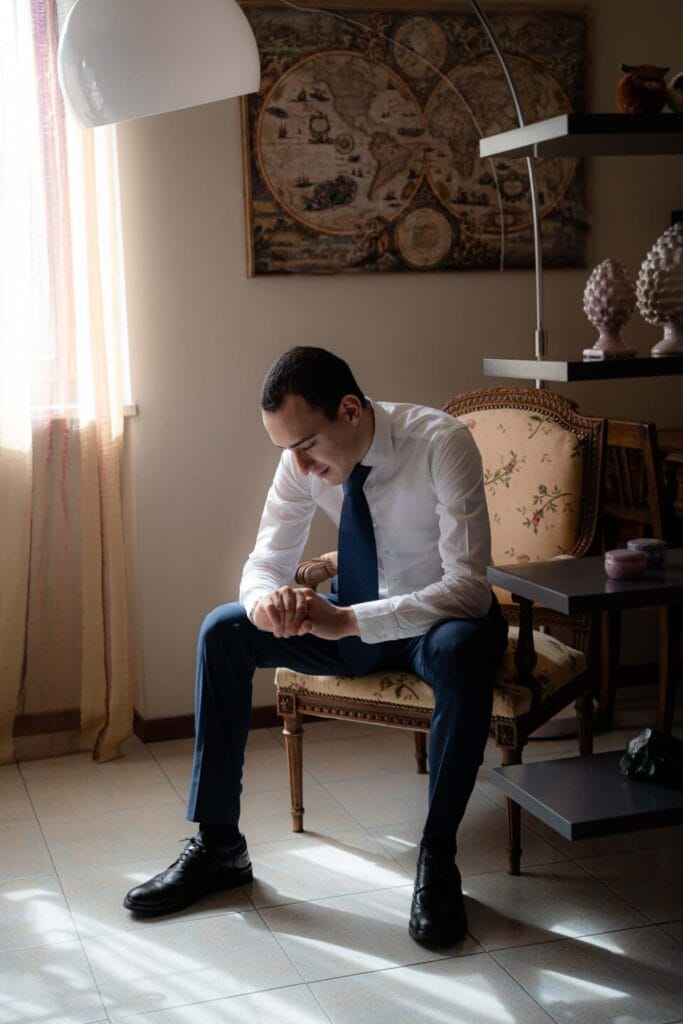 foto di dettaglio dello sposo seduto su una poltrona a casa mentre fissa il suo orologio, indossa pantaloni blu e una camicia bianca, vicino ad una finestra e sotto una lampada ad arco, durante servizio fotografico di matrmonio di yelley photography