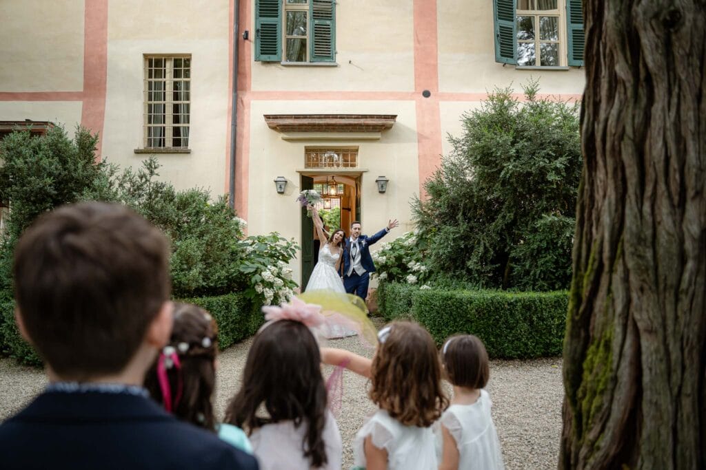 Foto dell'entrate degli sposi in location accolta dagli ospiti con nastri colorati, gli sposi escono da una porta di legno e ci sono dei bambini in primo piano, la timeline di matrimonio comprendeva questa fase quando è stata scritta
