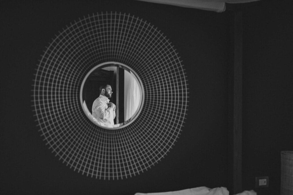 dettaglio dello sposo durante i preparativi in bianco e nero, la sua sagoma si vede da lontano dentro uno specchio circolare che sembra un sole ed è l'unico elemento chiaro della foto durante servizio fotografico di matrimonio di yelley photography