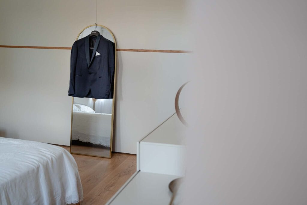 Bride and groom's jacket hanging in the distance near a mirror, seen in passing as an outside observer, the jacket is dark blue and the room is light and appropriate for the bride and groom's preparations during wedding