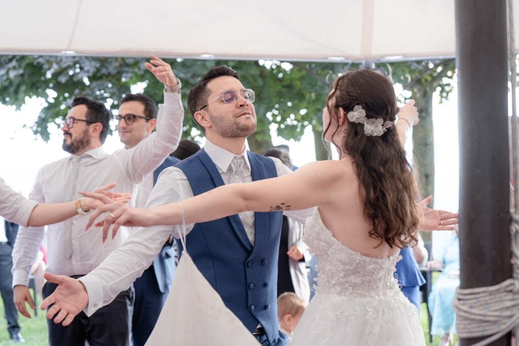 Sposi che ballano felici guardandosi negli occhi durante servizio di matrimonio, vestiti di bianco lei e di blu lui, intorno hanno tante persone che ballano
