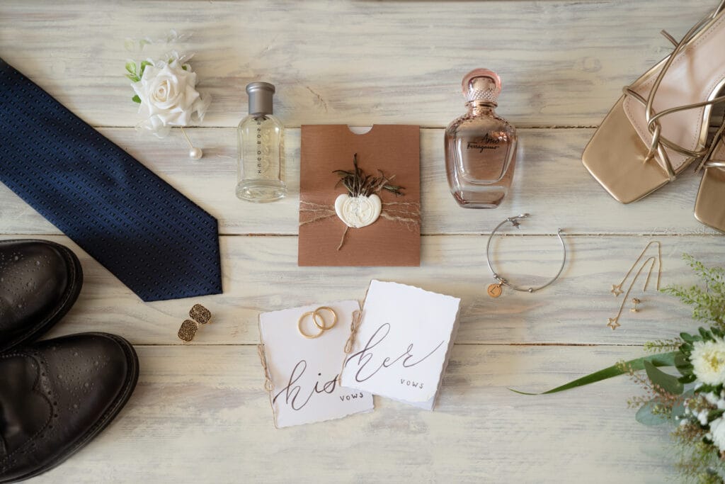 Foto dei dettagli dei preparativi degli sposi con libretti delle promesse di matrimonio, scarpe degli sposi, profumi e gioielli sparsi su un tavolo di legno visto dall'alto della fotografa yelley photography