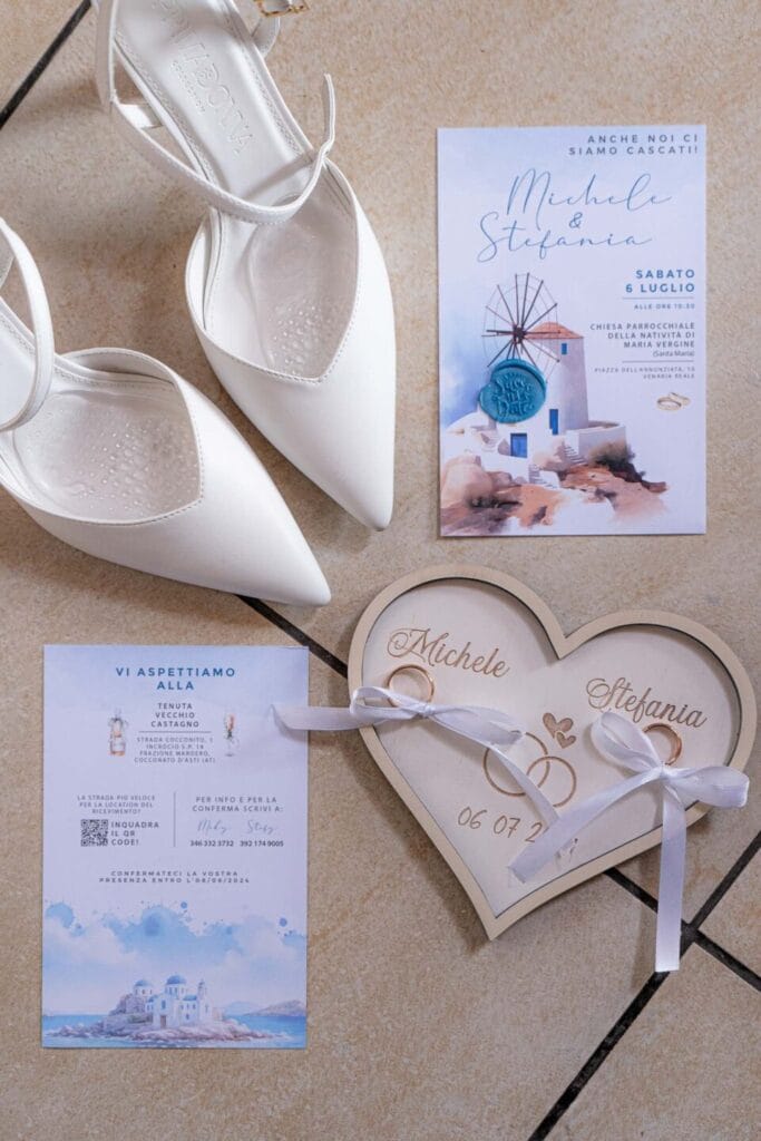 Foto delle scarpe della sposa, anelli e partecipazioni di matrimonio disposti in ordine sul pavimento, le scarpe sono a punta bianche, il portafedi è un cuoricino di legno e le partecipazioni hanno lo stile marino con un faro tutto azzurro