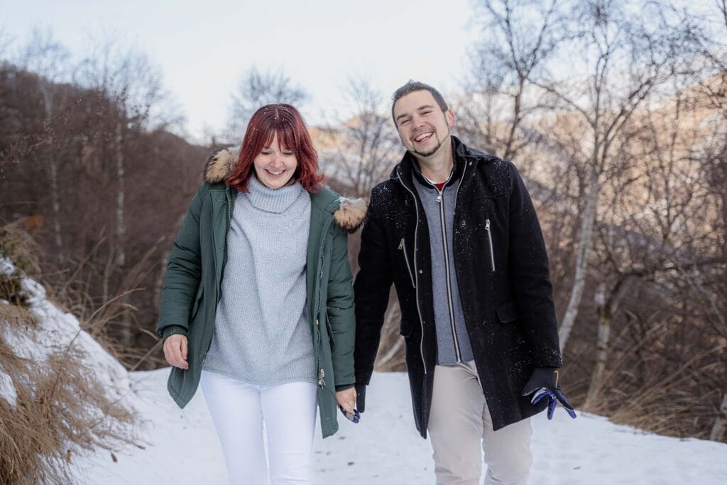 Coopia che passeggia in mezzo alla neve con indosso giacconi verde e nero, maglione celeste e pantaloni chiari, sorridono e hanno alle spalle degli alberi
