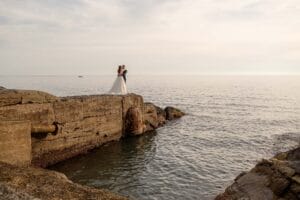 foto di coppia al mare in liguria a camogli durante servizio fotografico di matrimonio, sono sopra uno scoglio in mezzo al mare, visibili in lontananza con la luce del tramondo che li illumina