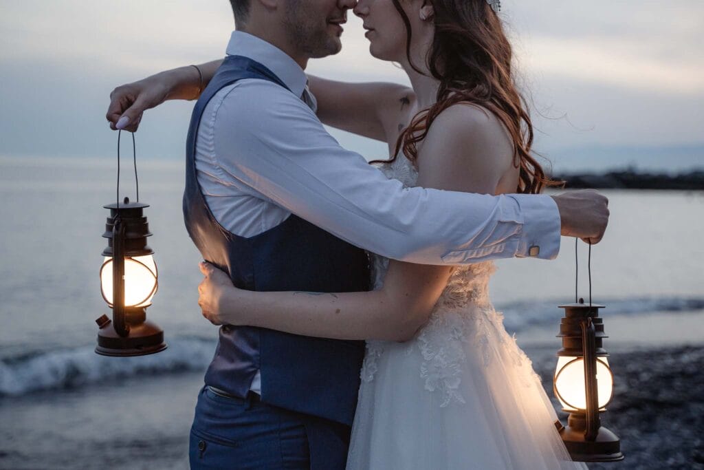 foto di una coppia durante prematrimoniale al mare in estate, che regge in mano due lanterne mentre si abbracciano indossando gli abiti da sposi, nello sfondo si vede il mare al tramonto di Camogli, in Liguria, scattato durante servizio prematrimoniale di yelley photography